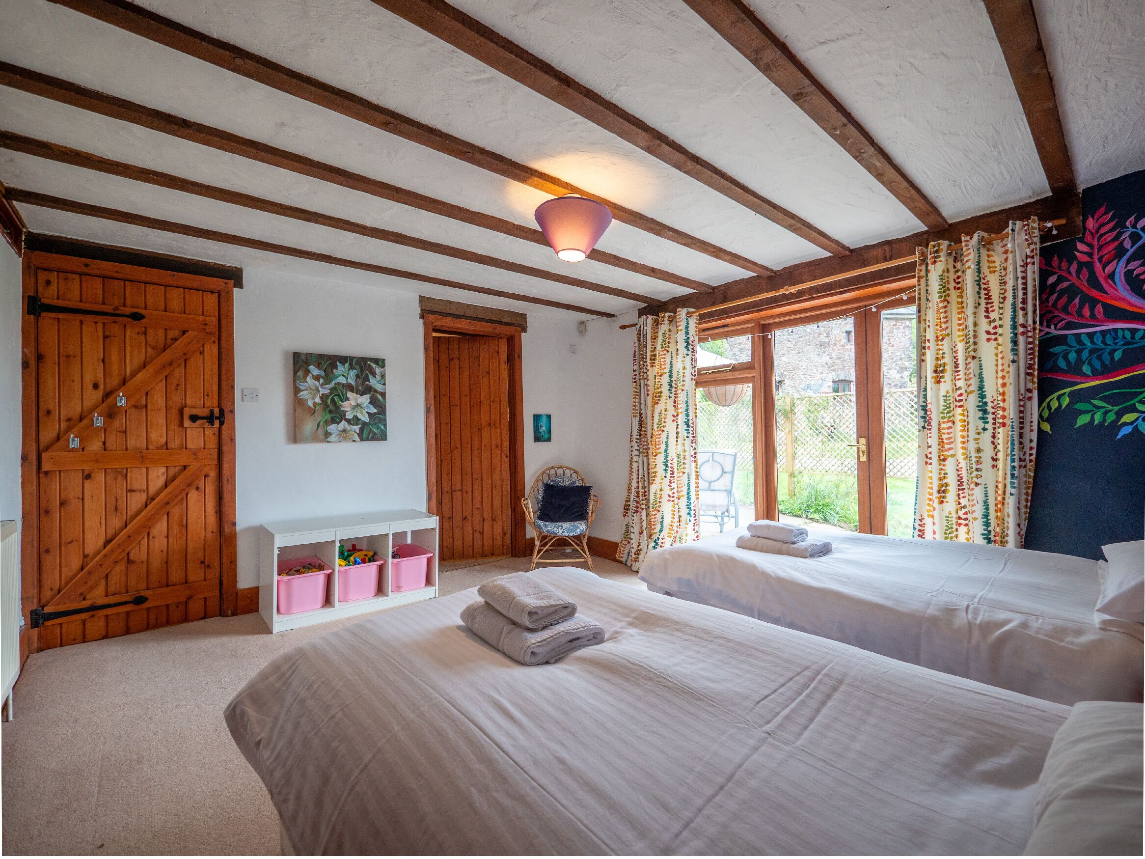 A bedroom with two beds, wooden beams, a ceiling light, a window with curtains, a chair, and a wall-mounted shelf with storage bins.