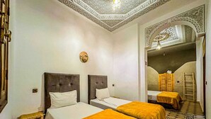 A hotel room with two beds, a small wooden stool, a decorative wall hanging, and a patterned floor. - Riad Malayine Sarl (Marrakech)