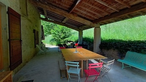 An outdoor covered patio with a wooden table and chairs, a bench, and a view of greenery. - La ferme Biscot : Charm & Nature 45 min from Lyon. Renovated farmhouse with swimming pool (Ambronay)