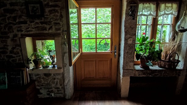 Une porte en bois donne sur une pièce dont un mur est en pierre, une fenêtre offre une vue sur la verdure et une autre fenêtre est ornée d’un rideau.