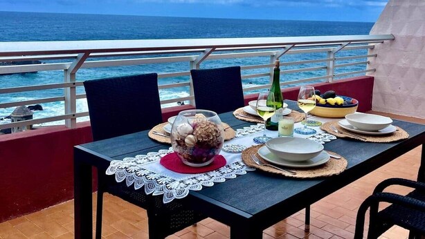 Spisebord dækket med tallerkener, et glas vin og en skål med frugt på en balkon med udsigt over havet.