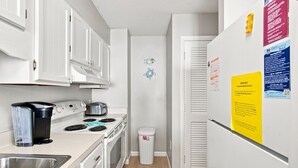 Een kleine keuken met witte kastjes, een fornuis, een spoelbak en een afvalbak. Er zijn bordjes op de koelkast en een kleine visdecoratie aan de muur.