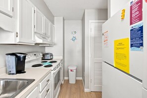 A compact kitchen with white cabinets, a stove, a sink, and a trash bin. There are signs on the refrigerator and a small fish decoration on the wall. - Beautifully Upgraded!  Pool and Beach Access (North Myrtle Beach)