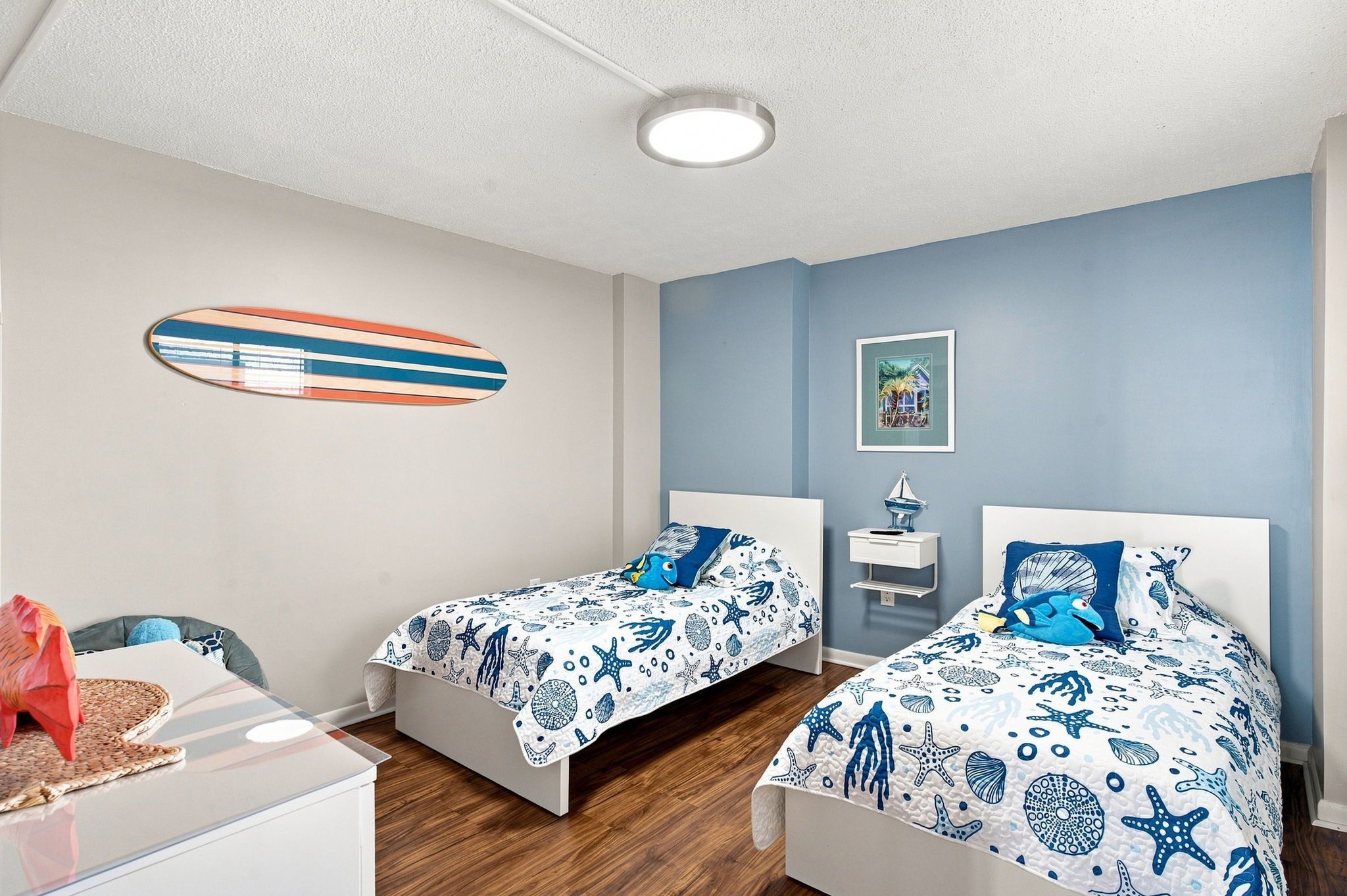 Ein Schlafzimmer mit zwei Betten, einer Surfbrettermalerei, einem gerahmten Bild und einem kleinen Tisch mit einer Lampe.