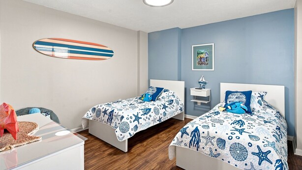Een slaapkamer met twee bedden, een decoratie van een surfplank, een lijst met een foto en een klein tafeltje met een lamp.