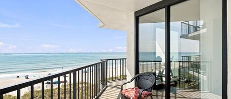 Un balcon donnant sur la plage et l’océan, une chaise et une porte vitrée qui mène à un espace intérieur.