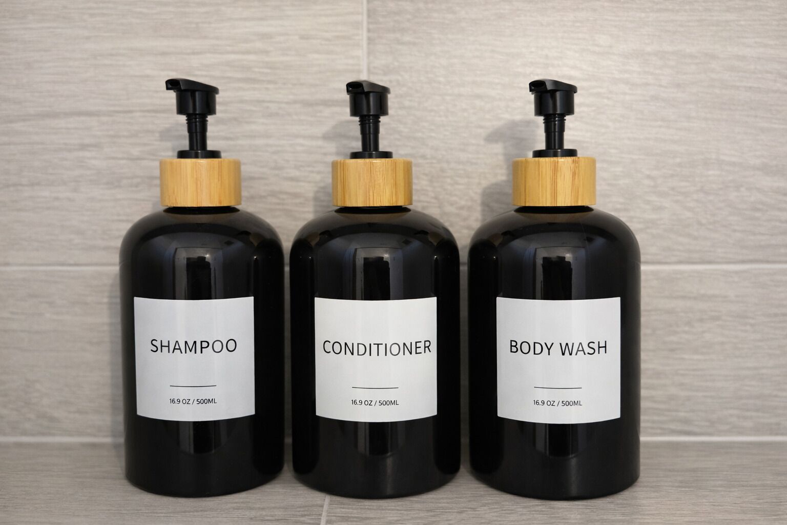 Three black bottles with wooden pumps and labels, each labeled as shampoo, conditioner, and body wash.