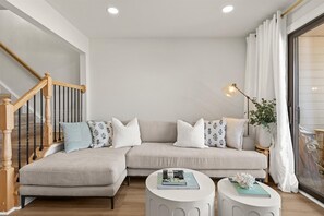 Ein Wohnzimmer mit einem beigefarbenen Ecksofa, zwei weißen runden Beistelltischen, einer Holztreppe und einem großen Fenster mit weißen Vorhängen.