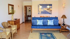 Een woonkamer met een blauwe bank, rieten stoelen, een houten kast, een schilderij en een lamp.