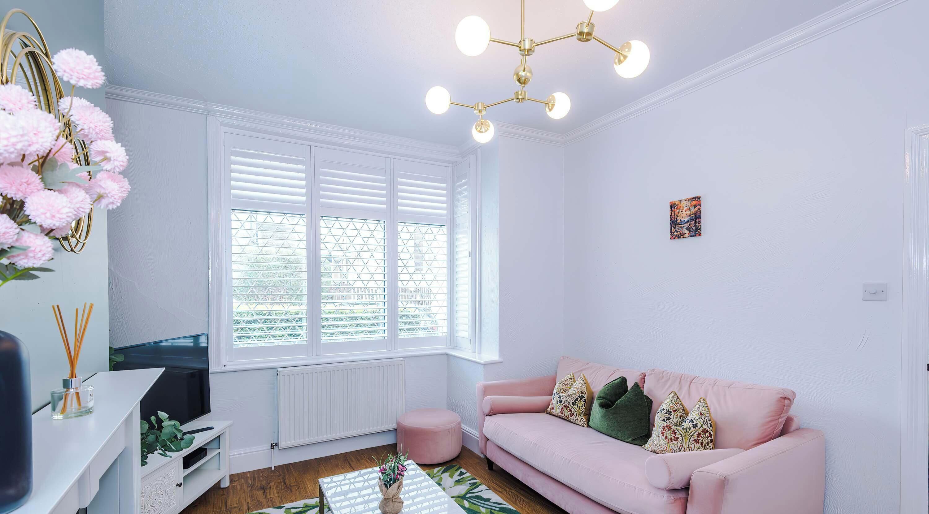 Una sala de estar con un sofá rosa, un radiador blanco, una ventana con persianas, una araña y una pequeña obra de arte en la pared.
