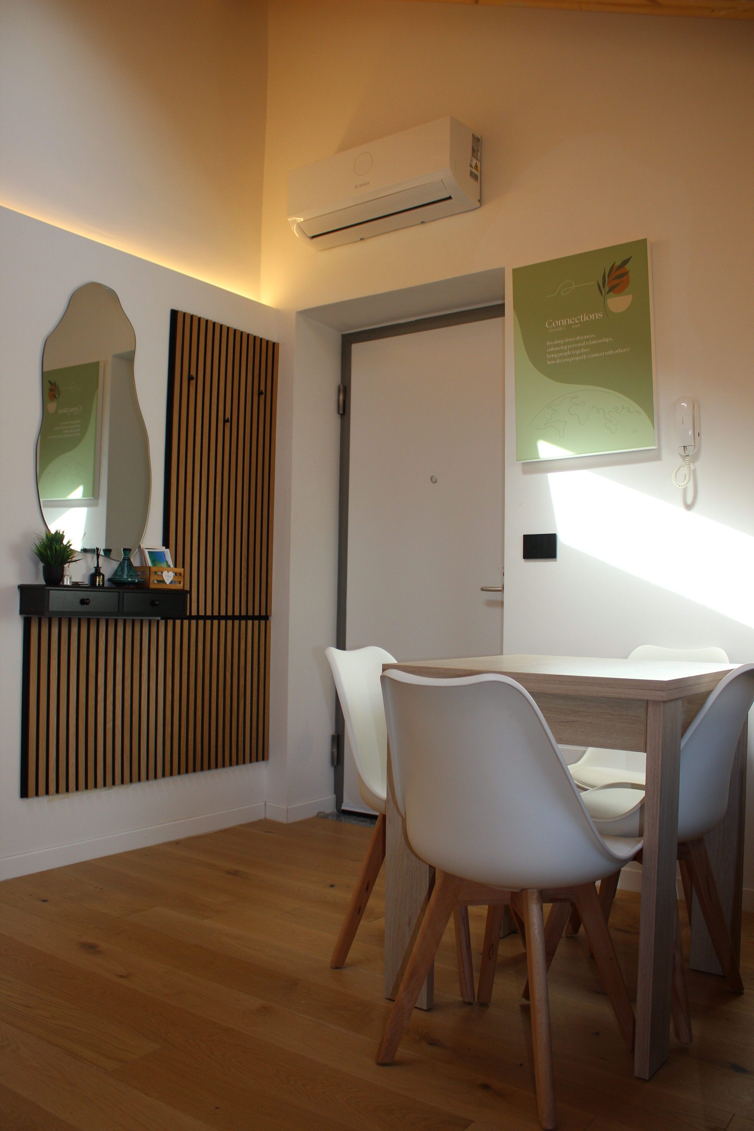A modern room with a wooden floor, a white table with chairs, a wall-mounted air conditioner, a framed picture, and a decorative panel.