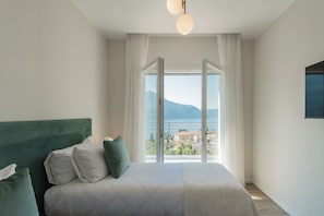 Una camera da letto con un letto, vista su un corpo d'acqua e edifici, e una finestra con tende.