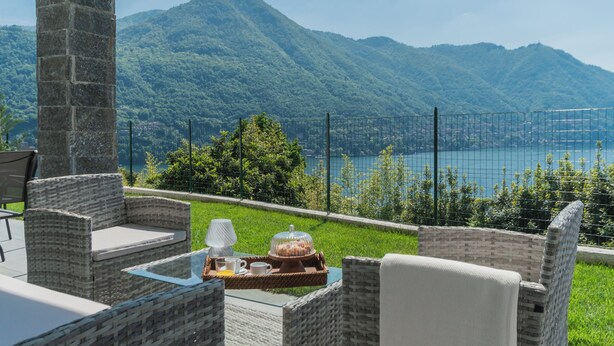Eine Terrasse mit Korbmöbeln, einem Glastisch und Blick auf Berge und einen See.