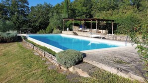 A rectangular swimming pool with a wooden deck and lounge chairs, surrounded by a stone patio and lush greenery.
