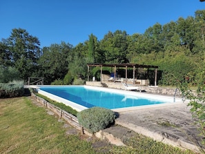 Una piscina rettangolare con un prendisole di legno e poltrone a sdraio, circondata da un patio in pietra e vegetazione lussureggiante.