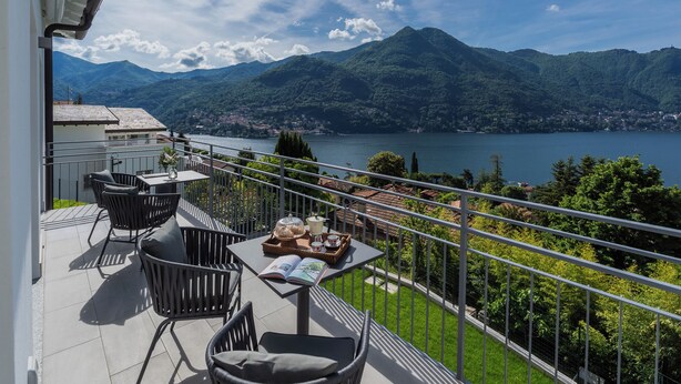 Un balcon aménagé avec du mobilier d’extérieur, une table avec un livre et un plateau rempli d’objets, et une vue sur un lac et des montagnes.