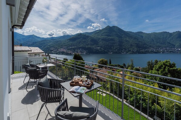 Un balcon aménagé avec du mobilier d’extérieur, une table avec un livre et un plateau rempli d’objets, et une vue sur un lac et des montagnes.