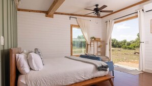 A bedroom with a bed, wooden beams, a ceiling fan, and a view of the outdoors. - Cedar Haus - king bed and soaking tub (Marble Falls)