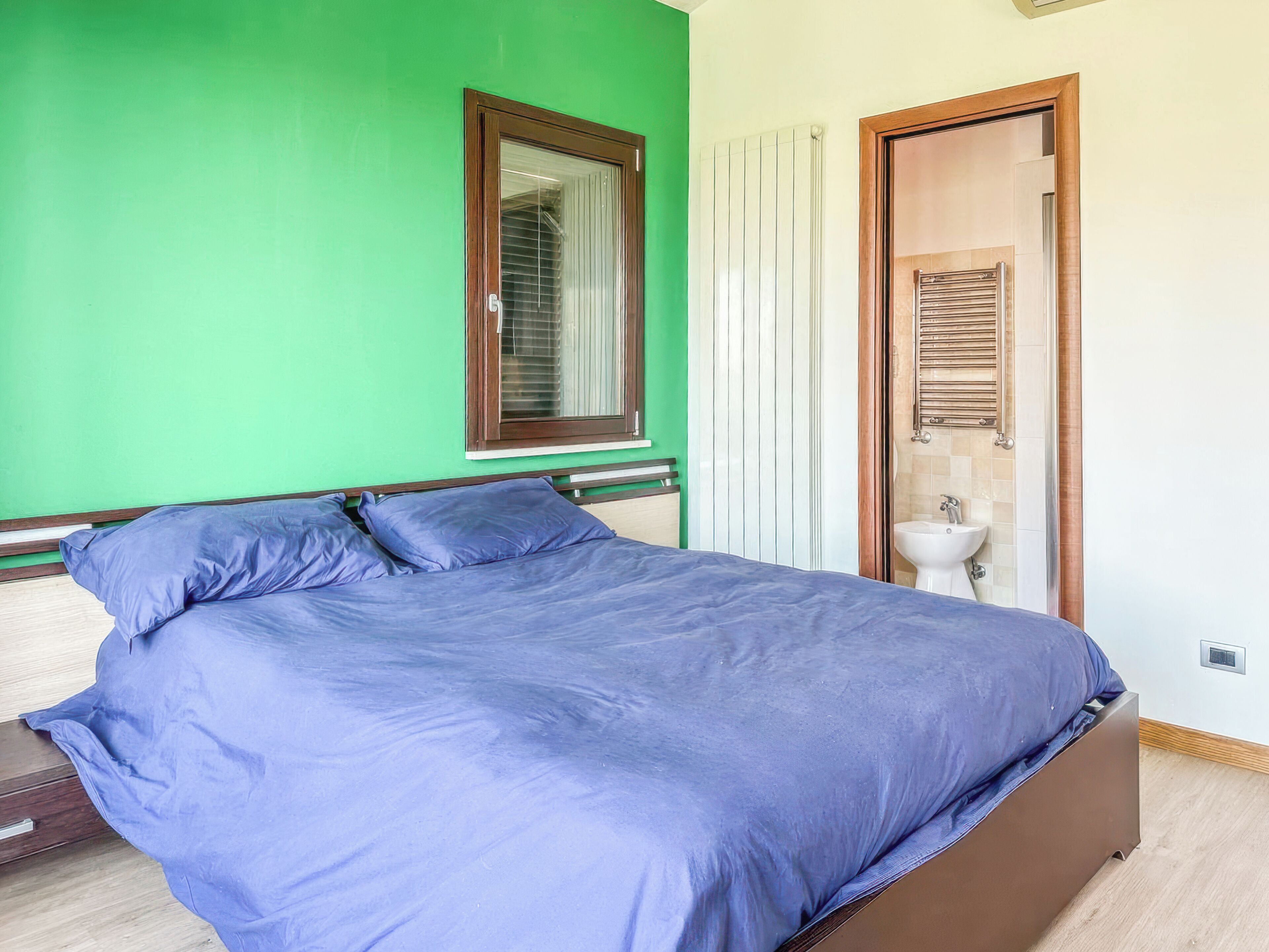 Une chambre à coucher avec un lit, une fenêtre avec des stores, un radiateur et une porte donnant sur une salle de bain.