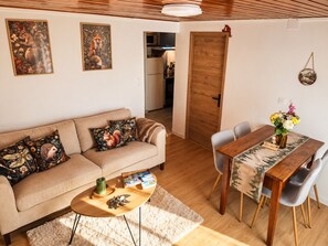 Ein gemütliches Wohnzimmer mit einer Couch, einem Couchtisch und einem Esstisch mit Stühlen. An der Wand hängen zwei gerahmte Bilder, eine Holztür und im Hintergrund ist eine Küche zu sehen.