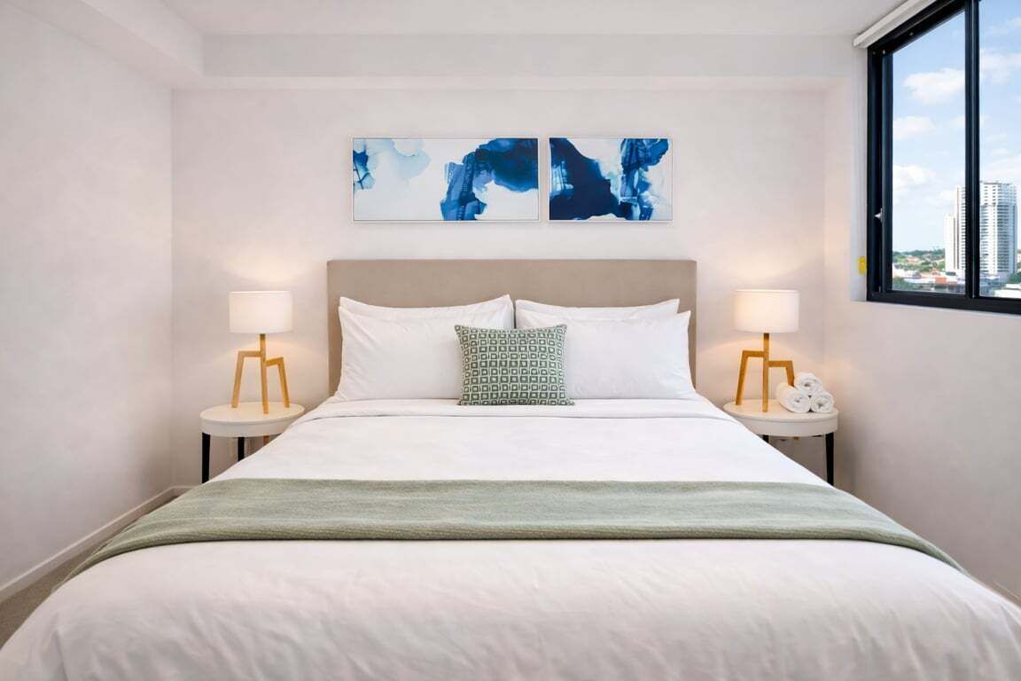 A hotel room with a bed, two bedside tables, a window with a city view, and abstract wall art.