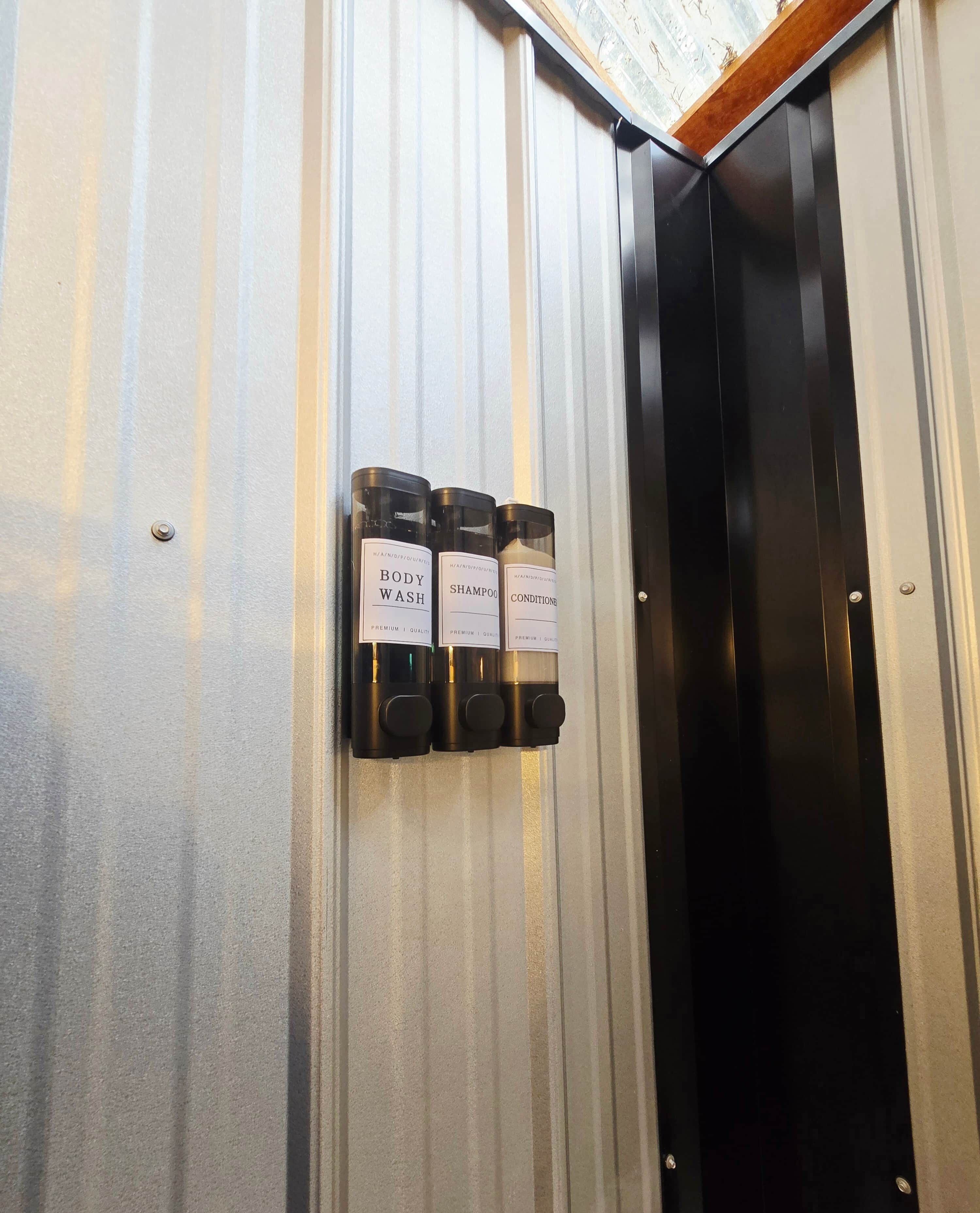 A wall-mounted dispenser with three bottles labeled 'BODY WASH,' 'SHAMPOO,' and 'CONDITIONER' in a hotel setting.