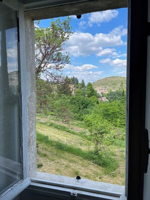 Ein Fenster mit Blick auf eine Grünfläche, Bäume und eine Hügelkette mit Gebäuden in der Ferne.