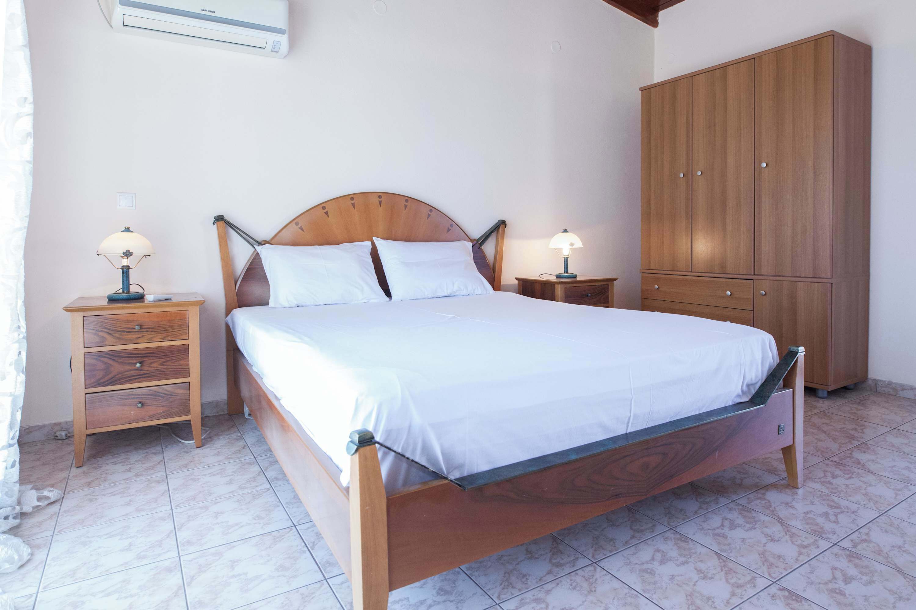 Ein Schlafzimmer mit einem hölzernen Bett, zwei Nachttischen mit Lampen, einem Kleiderschrank und einem Fliesenboden.