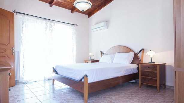 A bedroom with a wooden bed, a nightstand, a lamp, a window with curtains, and an air conditioning unit. - Peristeri 1 Bd Apt w Balcony and View (Peristeri)