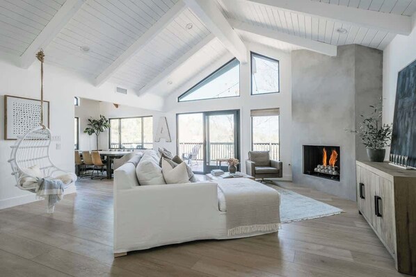 A spacious living room with a high ceiling, large windows, a fireplace, and a dining area. - The Boujee Basecamp, by Tag Home Design (Lake Arrowhead)