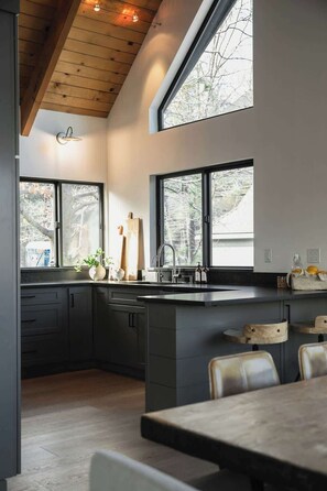 A modern kitchen with dark cabinetry, a large island, and a dining area with a table and chairs. - The Boujee Basecamp, by Tag Home Design (Lake Arrowhead)