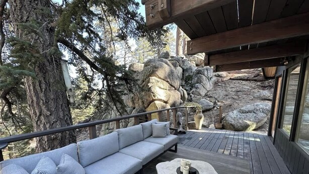 Une terrasse en bois avec un canapé gris et une table en pierre, entourée de gros rochers et d’arbres.
