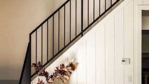 Una escalera con barandilla negra, pared blanca, sofá blanco con manta beige y una mesita con un jarrón blanco y una lámpara.
