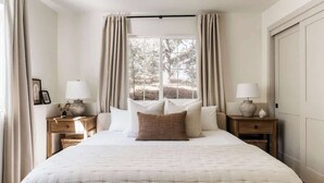 Un dormitorio con una cama grande, dos mesitas de noche con lámparas, un ventanal con cortinas y un banco de madera.