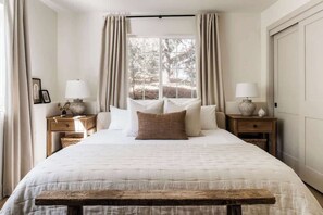 A bedroom with a large bed, two bedside tables with lamps, a window with curtains, and a wooden bench. - Elmwood Lake House, by @halfway_wholeistic (Lake Arrowhead)