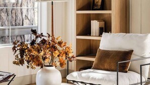 Una sala moderna con un ventanal grande, un sofá blanco, una estantería de madera y un jarrón decorativo con flores secas.