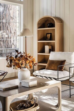 A modern living room with a large window, a white sofa, a wooden bookshelf, and a decorative vase with dried flowers. - Elmwood Lake House, by @halfway_wholeistic (Lake Arrowhead)