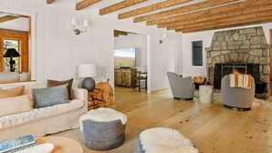 Een ruime woonkamer met een open haard, houten balken, een bank, poefs en een eethoek.
