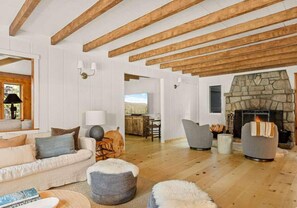 Ein geräumiges Wohnzimmer mit Kamin, Holzbalken, einer Couch, Poufs und einem Essbereich.