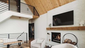 Una sala de estar moderna con techo de madera, chimenea, televisor montado en la pared y escalera que sube a un nivel superior.