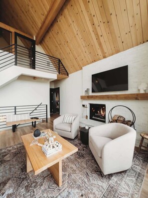 A modern living room with a wooden ceiling, a fireplace, a TV mounted on the wall, and a staircase leading to an upper level. - The Lakeview House (w/ hot tub, by @To_Dwell_Here) (Lake Arrowhead)