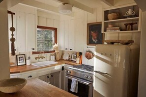 Una cocina con armarios blancos, encimera de madera, estufa, refrigerador y ventana con vistas a follaje.