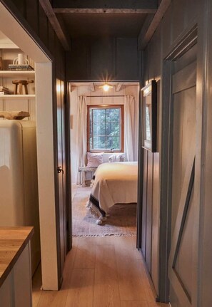 Un pasillo estrecho que conduce a un dormitorio con ventana, cama y una mesita.