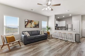 A modern living room with a sofa, a dining area with stools, a kitchen with white cabinets, and a ceiling fan.