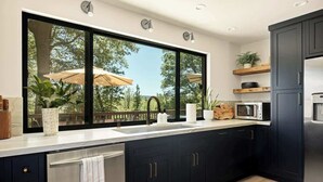 A modern kitchen with dark cabinets, a stainless steel dishwasher, a microwave, and a large window overlooking a deck with umbrellas. - Geneva Pines (walk to lake!) (Lake Arrowhead)