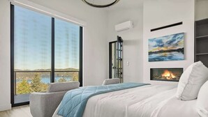A bedroom with a large window, a bed, a fireplace, and a painting on the wall.