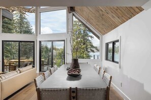 A modern dining room with a long table, cushioned seating, and large windows offering a view of trees. - Emerald Point Retreat, lakefront with dock! (Lake Arrowhead)