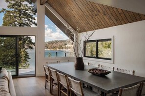 A dining area with a long table, chairs, a vase, and a view of a lake and trees. - Emerald Point Retreat, lakefront with dock! (Lake Arrowhead)