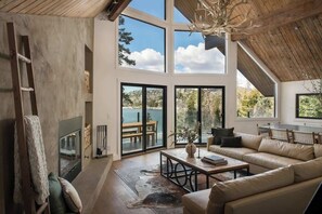 A spacious living area with a large window, a fireplace, a sofa, and a dining table. - Emerald Point Retreat, lakefront with dock! (Lake Arrowhead)