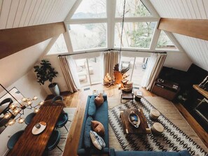 A spacious loft with a high ceiling, large windows, a dining area, a living room with a sofa, and a fireplace. - Harlow House, w/ hot tub, lake view & lake access! (Lake Arrowhead)